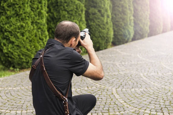 Profesionální svatební fotograf v černé košili a s ramenními popruhy v akci s obrázkem v zahradě. — Stock fotografie