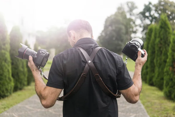 Fotograf profesjonalny wesele w czarnej koszulce i z ramiączkami, trzymając aparaty jak broń przeciwko zielony ogród. Fotografia ślubna. — Zdjęcie stockowe