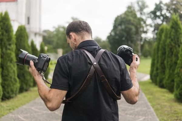 Fotograf profesjonalny wesele w czarnej koszulce i z ramiączkami, trzymając aparaty jak broń przeciwko zielony ogród. Fotografia ślubna. — Zdjęcie stockowe