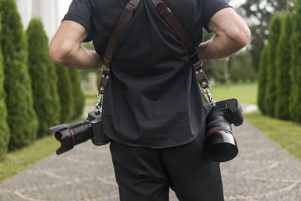 Back of professional wedding photographer in black shirt and with a two cameras an a shoulder straps against the green garden. Wedding photography.