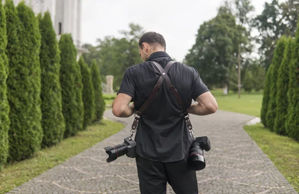 Back of professional wedding photographer in black shirt and with a two cameras an a shoulder straps against the green garden. Wedding photography.