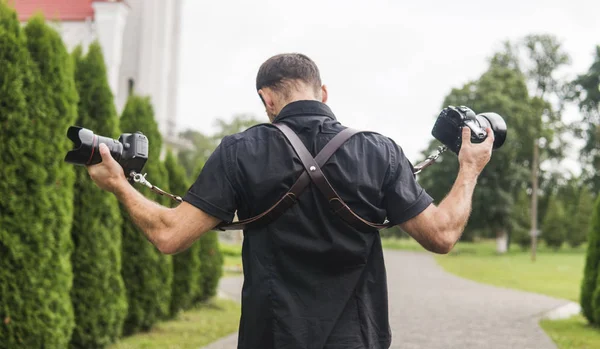 Profesionální svatební fotograf v černé košili a s ramínky držení kamery jako zbraně proti zelené zahrady. Svatební fotografie. — Stock fotografie