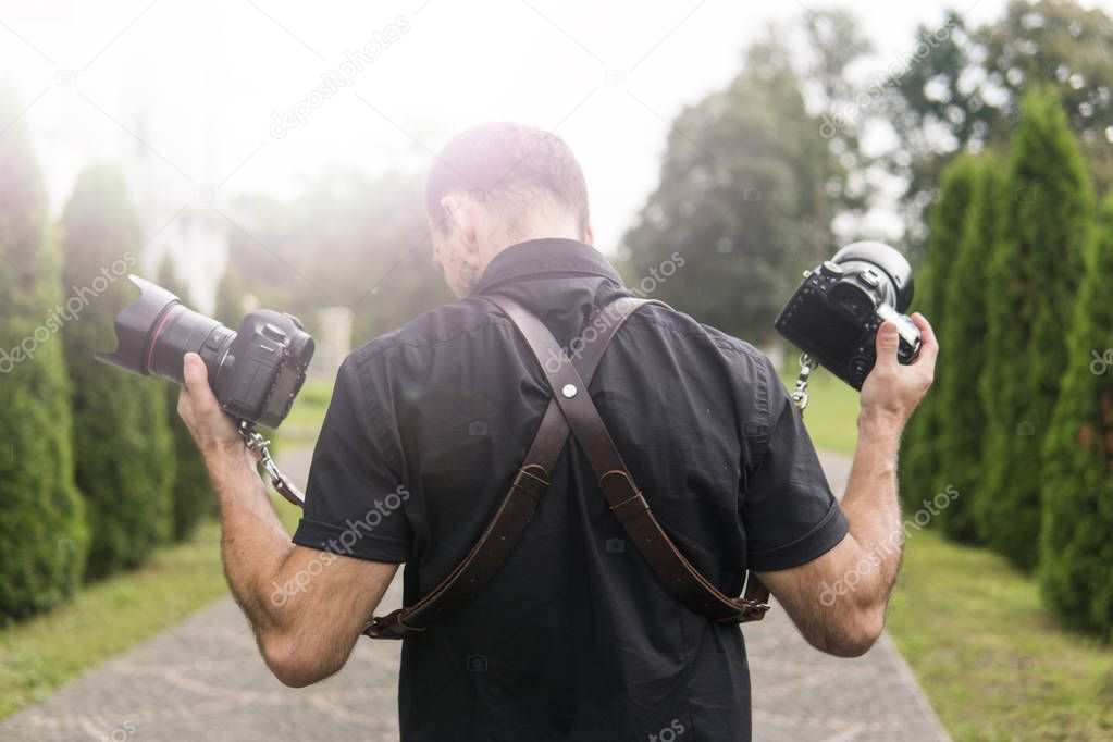 Professional wedding photographer in black shirt and with shoulder straps holding cameras like a guns against the green garden. Wedding photography.