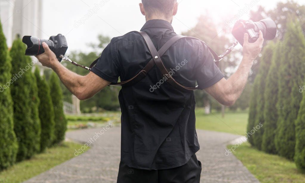 Professional wedding photographer in black shirt and with shoulder straps holding cameras like a guns against the green garden. Wedding photography.