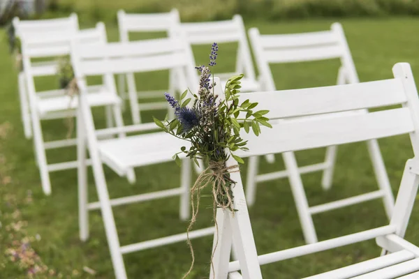 Witte bruiloft stoelen gedecoreerd met verse bloemen op een groen gras. Lege houten stoelen voor gasten op groene gazon in de tuin bereid voor huwelijksceremonie. — Stockfoto