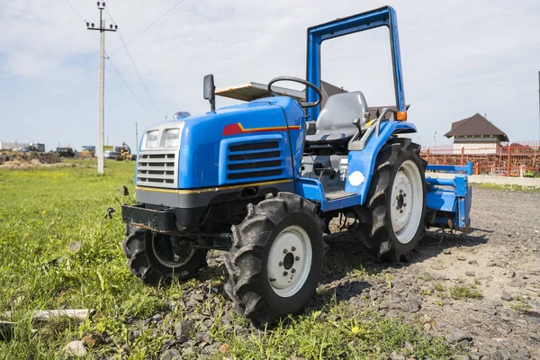A small mini blue tractor stands on a farm yard on green grass and waits for work to begin.