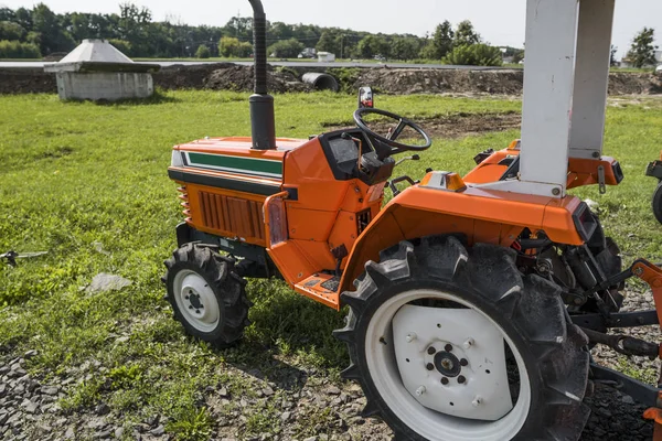 A small mini orange tractor stands on a farm yard on green grass and waits for work to begin.