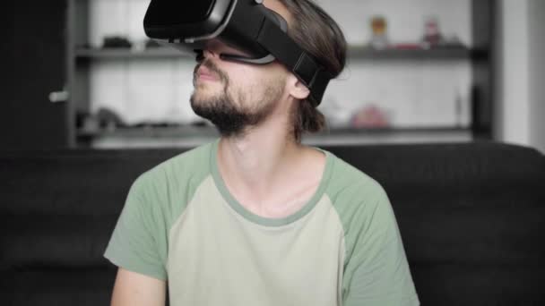 Молодой бородатый хипстер использует свой VR гарнитуру дисплей с наушниками для игры в виртуальную реальность или смотреть 360 видео, сидя на диване в гостиной. Технологии . — стоковое видео