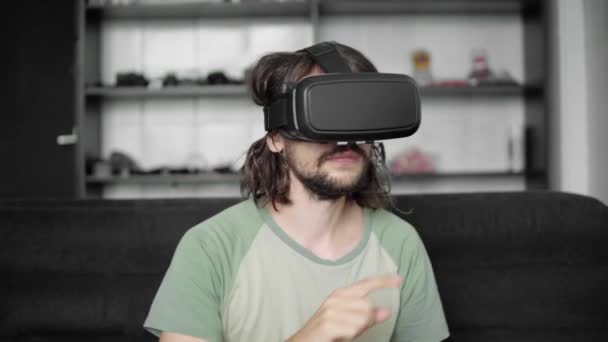 Молодой бородатый хипстер начинает использовать свой VR гарнитуру дисплей для навигации в vr-программе или игре виртуальной реальности, сидя дома на диване в гостиной. Технологии . — стоковое видео