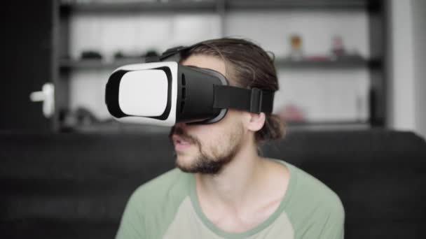 Молодой бородатый хипстер использует свой VR гарнитуру дисплей для игры в виртуальную реальность или смотреть 360 видео, сидя на диване в гостиной. Технологии . — стоковое видео