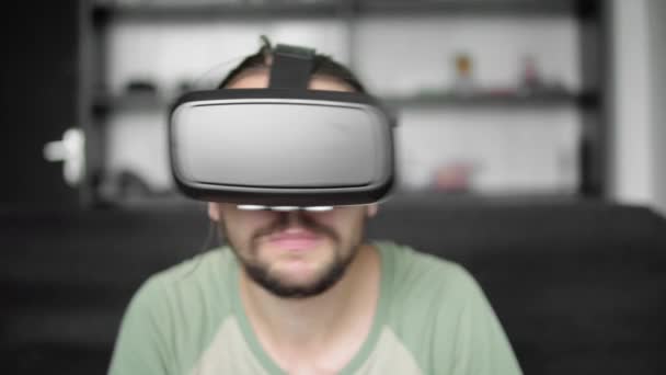 Молодой бородатый хипстер использует свой VR гарнитуру дисплей для просмотра 360 видео, сидя на диване и едят печенье дома в гостиной. Технологии . — стоковое видео