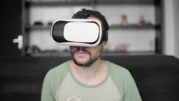 Jovem hipster barbudo usando sua tela de fone de ouvido VR para assistir ao vídeo 360 enquanto se senta no sofá e come biscoitos em casa na sala de estar. Tecnologia VR . — Vídeo de Stock