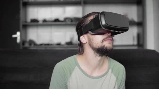Молодий бородатий hipster людина за допомогою його Vr гарнітура дисплея для віртуальної реальності гри або перегляду 360 відео, сидячи на дивані в домашніх умовах, у вітальні. VR технологій. — стокове відео
