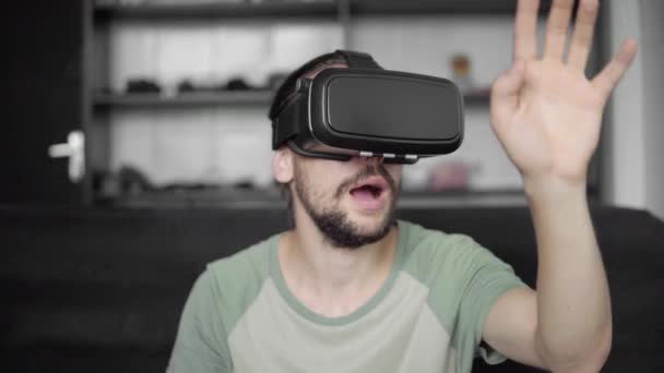 Fiatal szakállas csípő ember használ a Vr fejhallgató kijelző virtuális valóság játék, vagy néz a 360 video és kipróbálás-hoz érint-hoz valami úgy látja, a kanapén ülve. VR technológia. — Stock videók