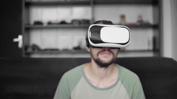 Giovane hipster barbuto che usa il suo visore VR per il gioco di realtà virtuale o guarda il video 360 e cerca di toccare qualcosa che vede seduto sul divano. Tecnologia VR . — Video Stock