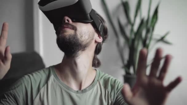 Unga skäggiga hipster man använder sin Vr headset display för virtuell verklighet spel eller tittar på en 360-video och försökte röra till något han se och fånga när du sitter på soffan. VR-tekniken. — Stockvideo