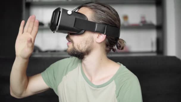 Fiatal szakállas csípő ember használ a Vr fejhallgató kijelző virtuális valóság játék, vagy néz a 360 video és kipróbálás-hoz érint-hoz valami úgy látja, a kanapén ülve. VR technológia. — Stock videók