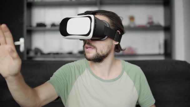 Joven hombre hipster barbudo utilizando su pantalla de auriculares VR para el juego de realidad virtual o ver el video 360 y tratando de tocar algo que ve mientras está sentado en el sofá. Tecnología VR . — Vídeo de stock