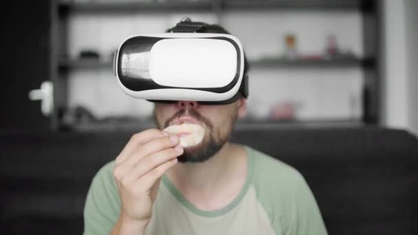 Молодой бородатый хипстер использует свой VR гарнитуру дисплей для просмотра 360 видео, сидя на диване и едят печенье дома в гостиной. Технологии . — стоковое видео