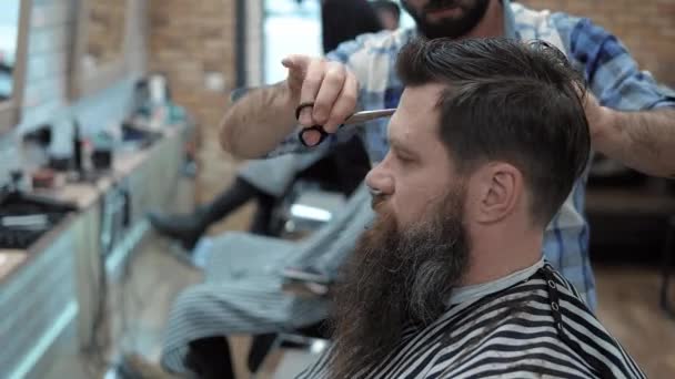 Close-up de barbeiro corta o cabelo por tesoura na barbearia. Cabeleireiros mãos no processo de trabalho. Barbeiro fazendo corte de cabelo de homem barbudo atraente na barbearia. Cabeleireiro no trabalho. Salão de beleza . — Vídeo de Stock
