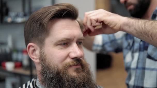 Close-up of barber cuts the hair by scissors at barbershop. Hairdressers hands at working process. Barber making haircut of attractive bearded man in barbershop. Hairdresser at work. Beauty saloon. — Stock Video