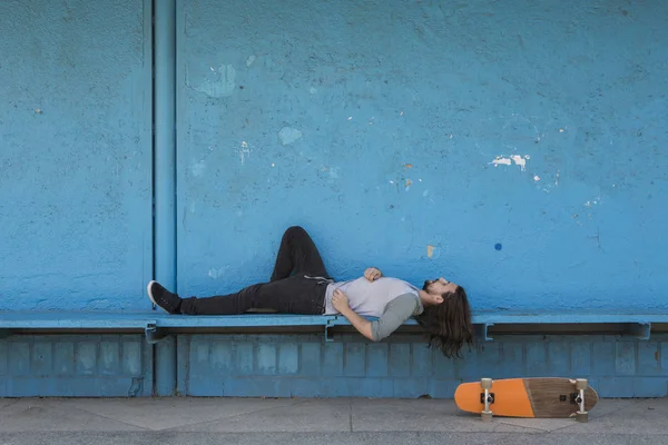 Скейтер лежит на синем фоне с оранжевым скейтбордом рядом с ним . Стоковая Картинка