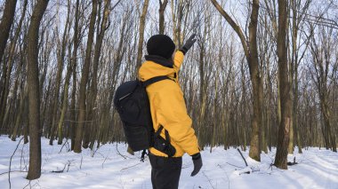 Adam sarı ceket ve siyah fotoğraf sırt çantası güneşli ormandaki bir ağaç görünümlü bir kar ile kaplı.