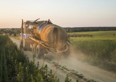 Bir buğday alanında bir yol boyunca bir tank ile mavi bir traktör sürmek.