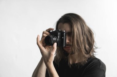 Yakışıklı genç sakallı adam uzun saçlı ve vintage eski moda film kamera beyaz bir arka plan üzerinde tutarak ve vizör kamera seyir siyah gömlekli.