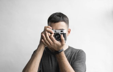 Sakallı yakışıklı genç bir siyah gömlekli vintage eski moda film kamera beyaz bir arka plan üzerinde tutarak ve kameranın vizör arıyorsunuz.