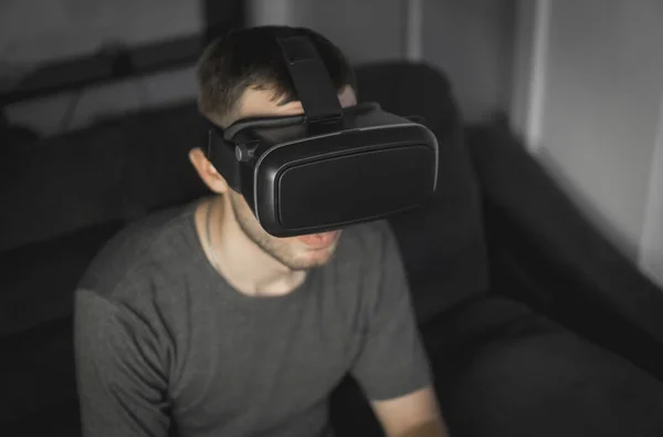Młody Brodaty mężczyzna noszenie zestawu słuchawkowego wirtualnej rzeczywistości w biurze, siedząc na kanapie. — Zdjęcie stockowe