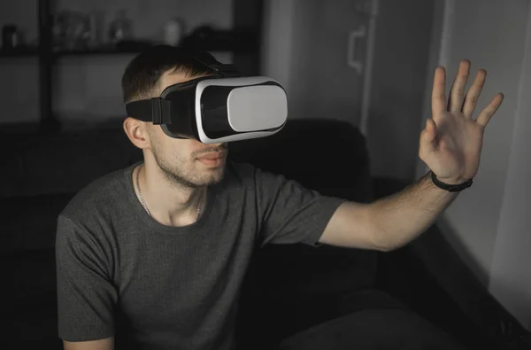 Young bearded man wearing headset of virtual reality in office sitting on a sofa and trying to touch something that he see in virtual world.