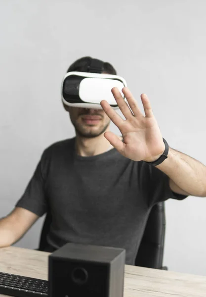 Jovem vestindo óculos de realidade virtual headset e sentado no escritório contra o computador. Conexão, tecnologia, nova geração. Homem tentando tocar objetos ou controlar VR com uma mão . — Fotografia de Stock