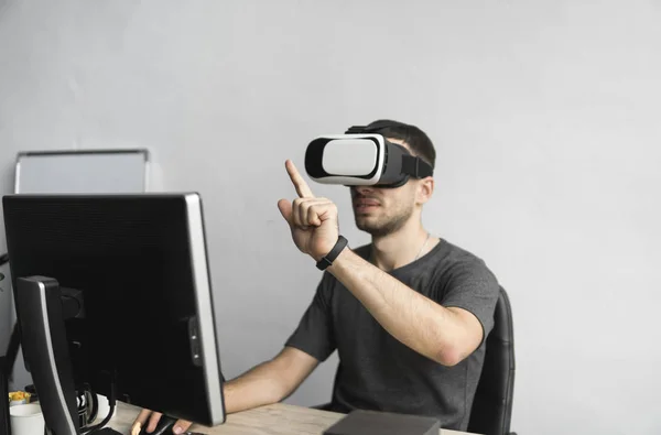Fiatal ember visel a virtuális valóság szemüveg, fejhallgató, és ülnek az irodában a számítógép ellen. Kapcsolat, technológia, új generációs. Ember próbál objektumok vagy vezérlőelem Vr egy kézzel érint. — Stock Fotó