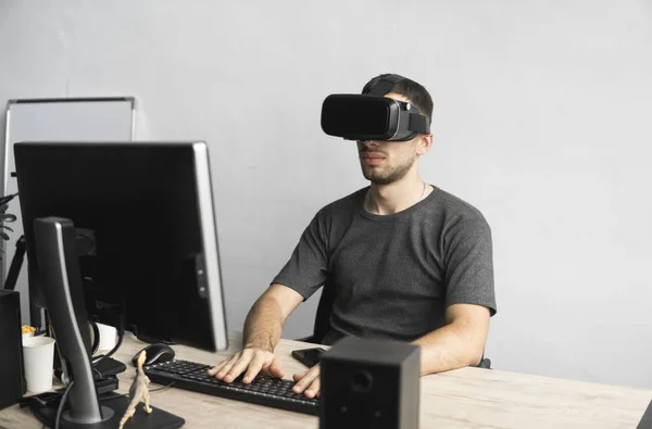 Νεαρός άνδρας φορώντας εικονική πραγματικότητα γυαλιά ακουστικό, vr κουτί και κάθεται στο γραφείο κατά την οθόνη του υπολογιστή. Σύνδεση, τεχνολογία, νέα γενιά, έννοια προόδου. — Φωτογραφία Αρχείου
