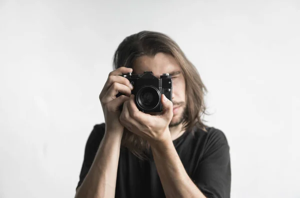 Bonito jovem barbudo homem com um cabelo longo e em uma camisa preta segurando câmera de filme antiquado vintage em um fundo branco e olhando no visor da câmera . — Fotografia de Stock