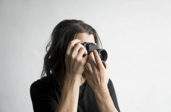 Bonito jovem barbudo homem com um cabelo longo e em uma camisa preta segurando câmera de filme antiquado vintage em um fundo branco e olhando no visor da câmera . — Fotografia de Stock