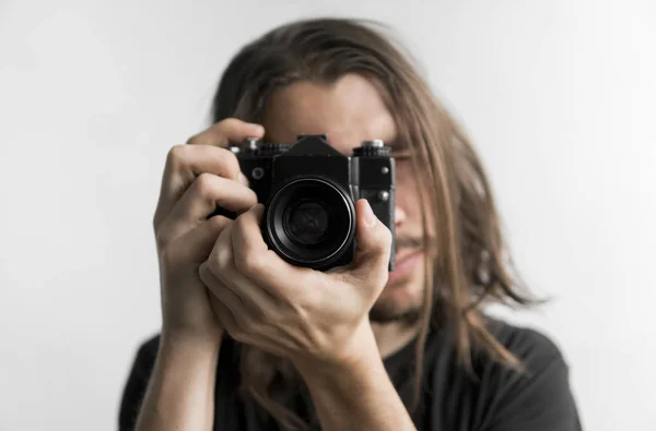 Hombre barbudo joven guapo con un pelo largo y una camisa negra que sostiene la cámara de cine antigua vintage sobre un fondo blanco y mirando en el visor de la cámara . —  Fotos de Stock