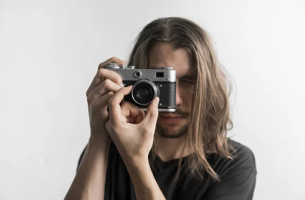 Hombre barbudo joven guapo con un pelo largo y una camisa negra que sostiene la cámara de cine antigua vintage sobre un fondo blanco y mirando en el visor de la cámara . —  Fotos de Stock
