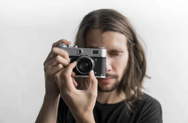 Bonito jovem barbudo homem com um cabelo longo e em uma camisa preta segurando câmera de filme antiquado vintage em um fundo branco e olhando no visor da câmera . — Fotografia de Stock