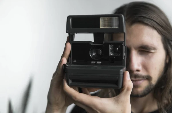 Bonito jovem barbudo homem com um cabelo longo e em uma camisa preta segurando câmera de filme antiquado vintage em um fundo branco e olhando no visor da câmera . — Fotografia de Stock