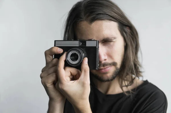 Bonito jovem barbudo homem com um cabelo longo e em uma camisa preta segurando câmera de filme antiquado vintage em um fundo branco e olhando no visor da câmera . — Fotografia de Stock