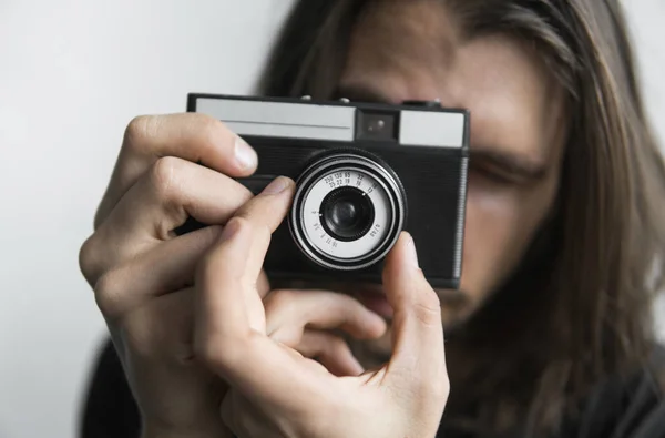 Pohledný mladý vousatý muž s dlouhými vlasy a v černé košili drží fotoaparát vintage staromódní film na bílém pozadí a při pohledu do fotoaparát hledáček. — Stock fotografie