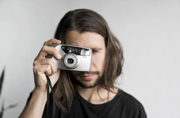 Bonito jovem barbudo homem com um cabelo longo e em uma camisa preta segurando câmera de filme antiquado vintage em um fundo branco e olhando no visor da câmera . — Fotografia de Stock