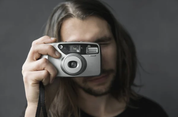 Bonito jovem barbudo homem com um cabelo longo e em uma camisa preta segurando câmera de filme antiquado vintage em um fundo preto e olhando no visor da câmera . — Fotografia de Stock