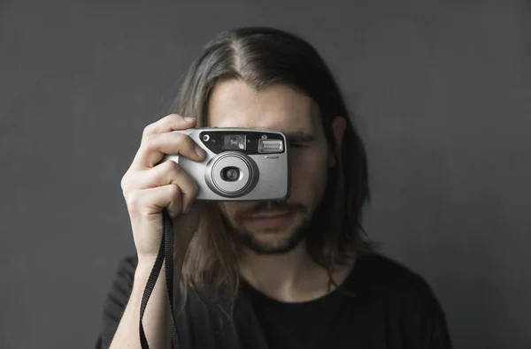 Hombre barbudo joven guapo con un pelo largo y con una camisa negra sosteniendo una cámara de película antigua vintage sobre un fondo negro y mirando en el visor de la cámara . —  Fotos de Stock
