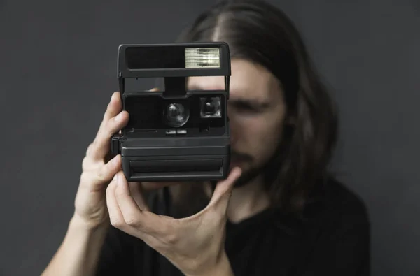 Bonito jovem barbudo homem com um cabelo longo e em uma camisa preta segurando câmera de filme antiquado vintage em um fundo preto e olhando no visor da câmera . — Fotografia de Stock