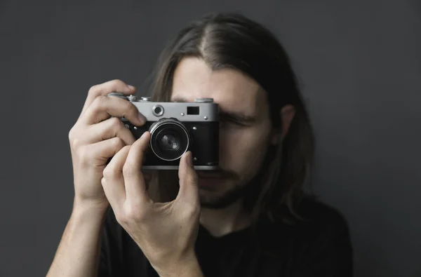 Bonito jovem barbudo homem com um cabelo longo e em uma camisa preta segurando câmera de filme antiquado vintage em um fundo preto e olhando no visor da câmera . — Fotografia de Stock