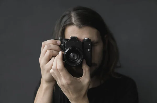 Handsome young bearded man with a long hair and in a black shirt holding vintage old-fashioned film camera on a black background and looking in camera viewfinder.