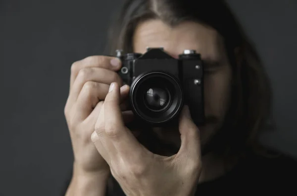 Knappe jonge bebaarde man met een lange haren en in een zwart shirt vintage ouderwetse filmcamera houden op een zwarte achtergrond en kijken in de camera zoeker. — Stockfoto